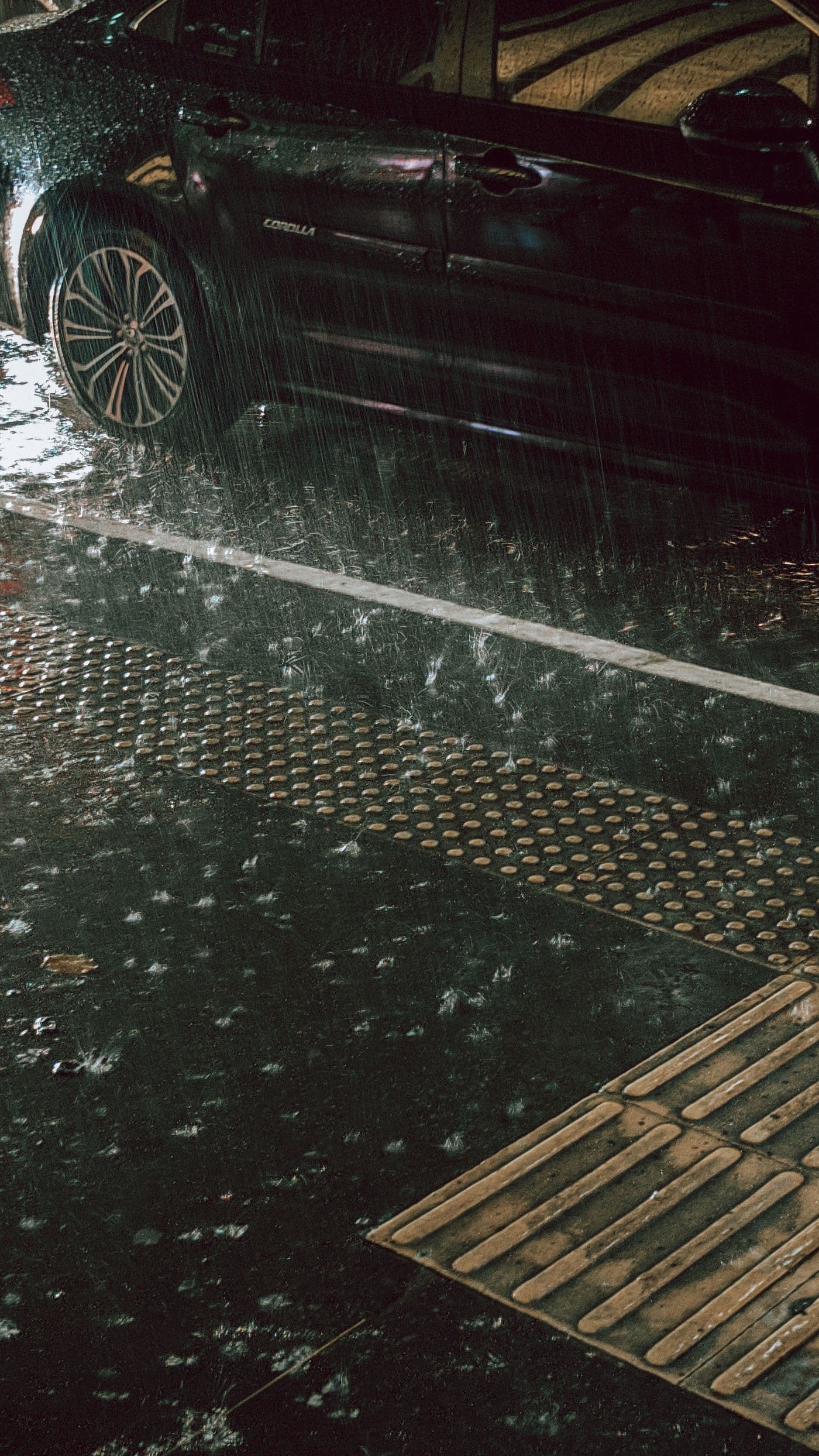 Un automóvil oscuro permanece junto a una acera mientras cae lluvia sobre la calzada.